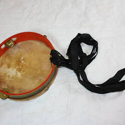 Tambourine frame underside is orange, natural animal skin. Metal jingles, bells and black ribbon streamers.