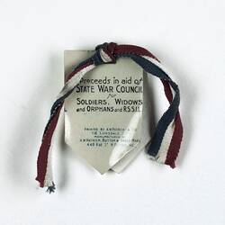 Celluloid-coated paper badge in shield shape with text and red, white and blue ribbon knotted at top.