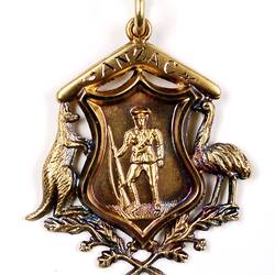 Gold ornament of Australian coat of arms with soldier holding rifle at centre, inscribed boomerang on top.