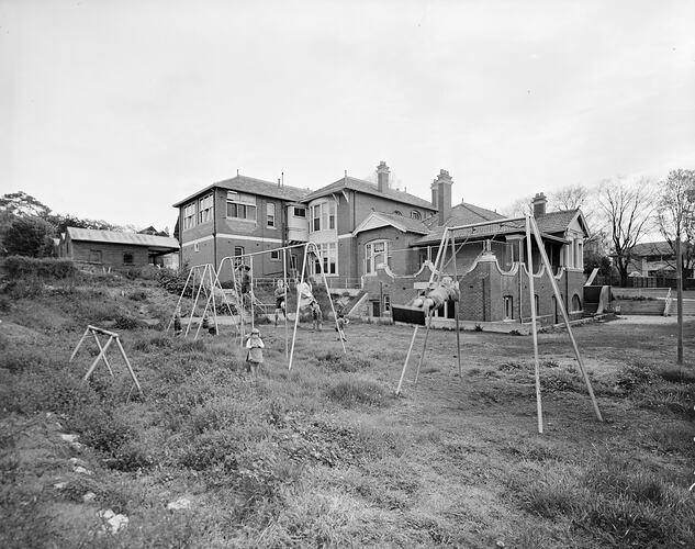 Children using playground equipment with a large house in the background.