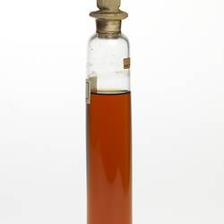 Cylindrical glass jar with brown liquid. Two labels affixed, sealed at top.