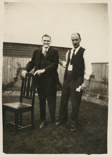 Young man standing in garden supported by a chair. An older man stands beside him, further supporting his arm.