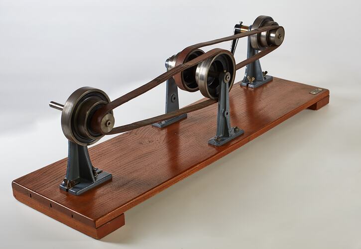 Wooden base with two pairs of round wheels, each connected via a pulley looped belt. One wheel has a handle.