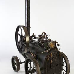 Model of black moveable steam engine on four wheels with tall chimney at front. Back view.