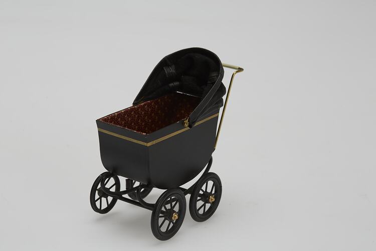 Painted black metal miniature pram with gold painted highlights and folding leather hood. Three quarter view.