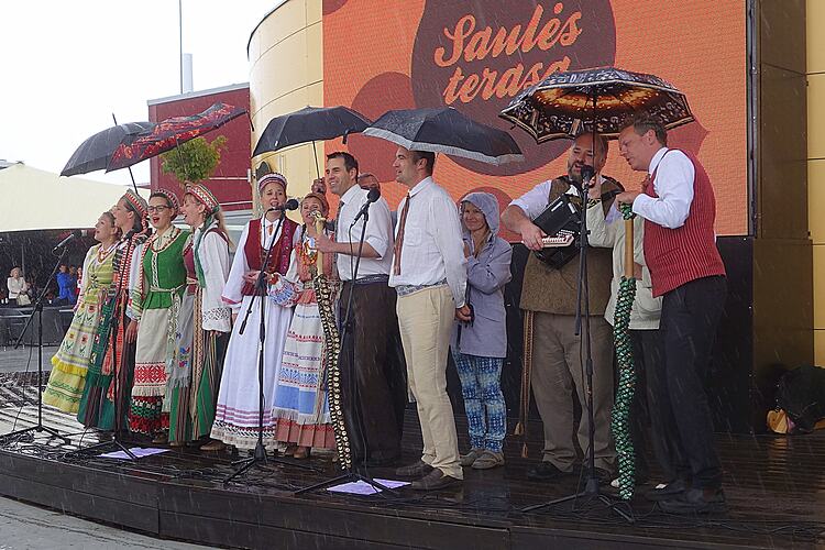 Ten people in Lithuanian costume perform under umbrellas. They sing, clap, play an accordion and two largerpho