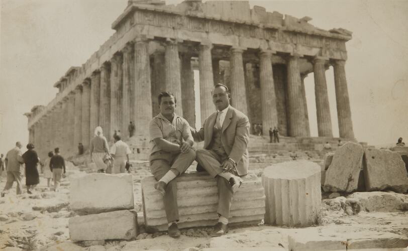 Two men sit on section of stone column with ancient ruins of columned Parthenon behind them.