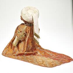 Back angled view of teddy bear wearing white turban, ornate embroidered coat with long tail.