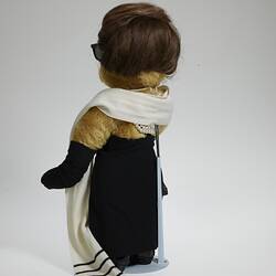 Back of light brown bear wears black hat, dress, full-length gloves, shoes and white scarf.