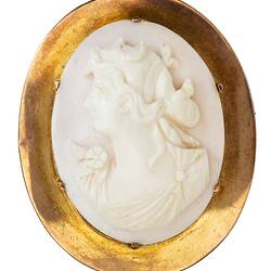 Oval white cameo of female bust facing left. She wears a draped garment and has ribbons in her hair. Plain gold setting.