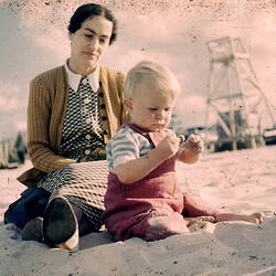 A woman sits on the beach with a blonde baby.