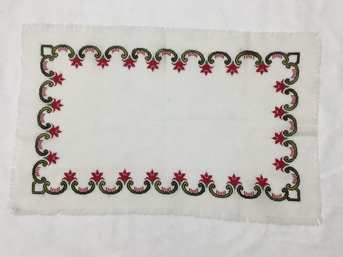 White rectangular cloth with border of green and red scalloped floral embroidery.