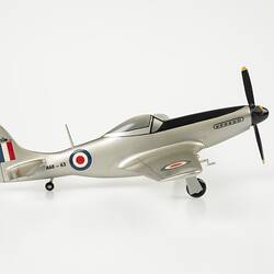 Wood model plane painted silver. Blue, white, red roundel on sides. Red, white, blue flag on tail. Front black four blade propeller. Three wheels. Profile.