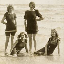 Four young women pose on beach shoreline wearing older style swimwear. Short sleeve tops and shorts.