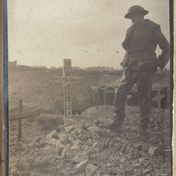 Soldier standing next to rock lined grave marked with white cross, other graves and shelter nearby.