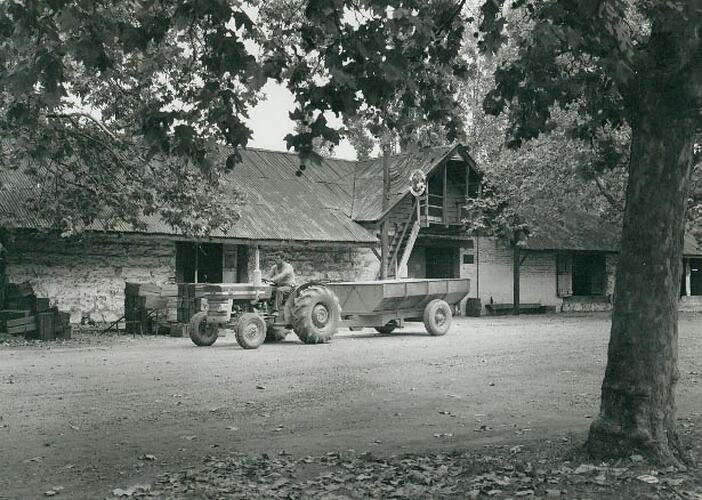 Man driving a tractor towing a farm trailer outside building.