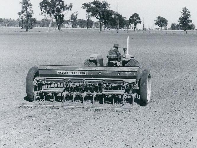 A man driving a tractor, pulling a seed drill with trailing small harrow.