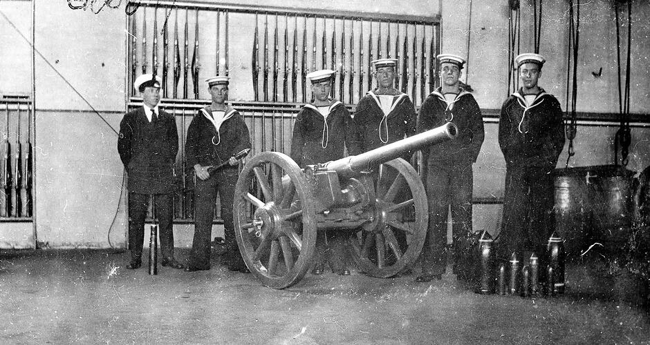 Row of sailors in an armoury with a field gun. Rifles in racks line the walls behind them.