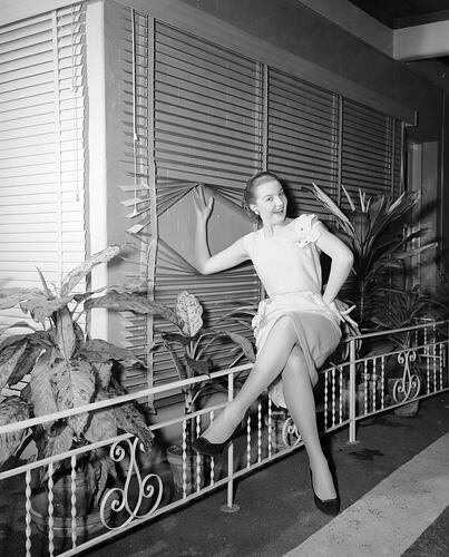 Woman perched on metal fence beside house and potplants. She smiles and holds open horizontal window blinds.