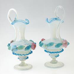 Pair of blue blown-glass ewer, spiral fluted neck, serpentine handle, flattened bulbous body layered with pink, blue, green flowers and leaves.