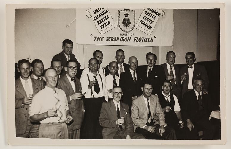 Group portrait of eighteen men in suits, with banner on wall behind.