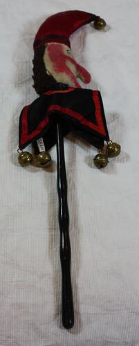 Mock sceptre with small jester's head made of cream cloth mounted on black wooden stick.