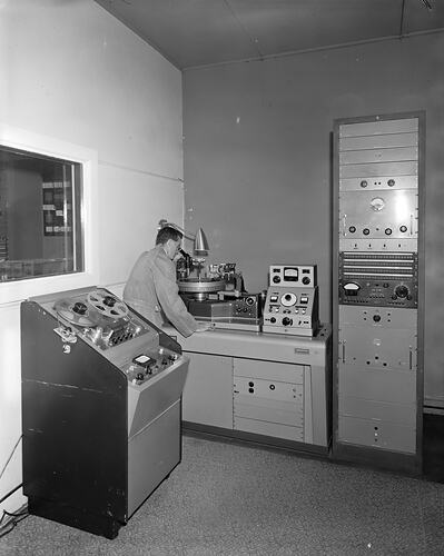 Man seen from behind looking into microscope at disc-shaped equipment. Consoles and cabinets surround him.
