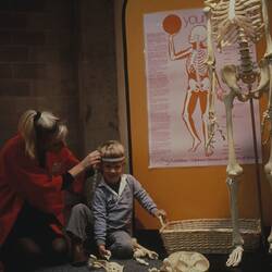 Woman measuring a smiling child's head. Poster and skeleton on display at the right. Yellow background.