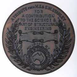 Round medal with raised text, blank scroll and Australia logo framed by wreath.