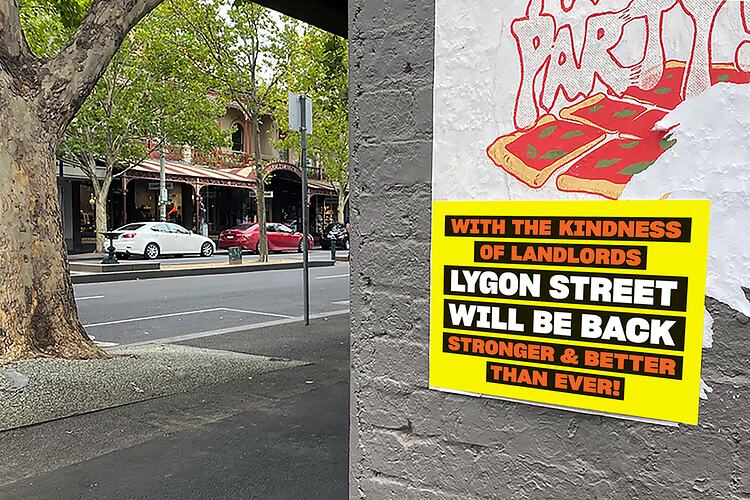 Yellow poster with red and white printed text. Affixed to edge of concrete wall. Streetscape in background.