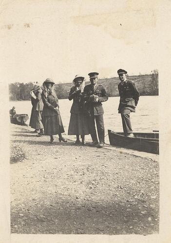 Three woman and two man standing by water, rowboats behind.