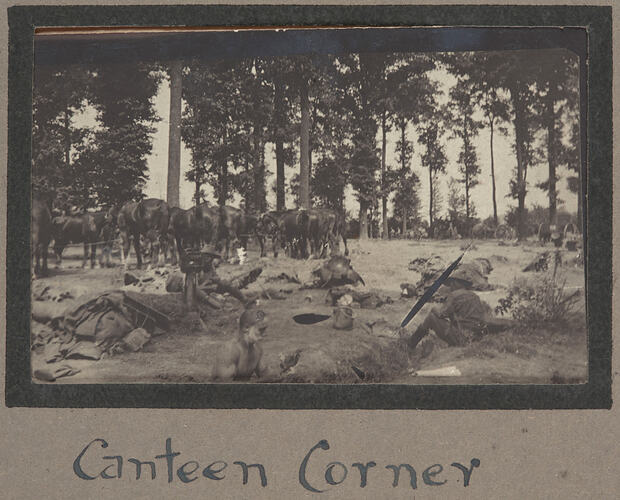 Servicemen resting in a camp in a tree lined field, in left mid ground is a group of unsaddled horses.