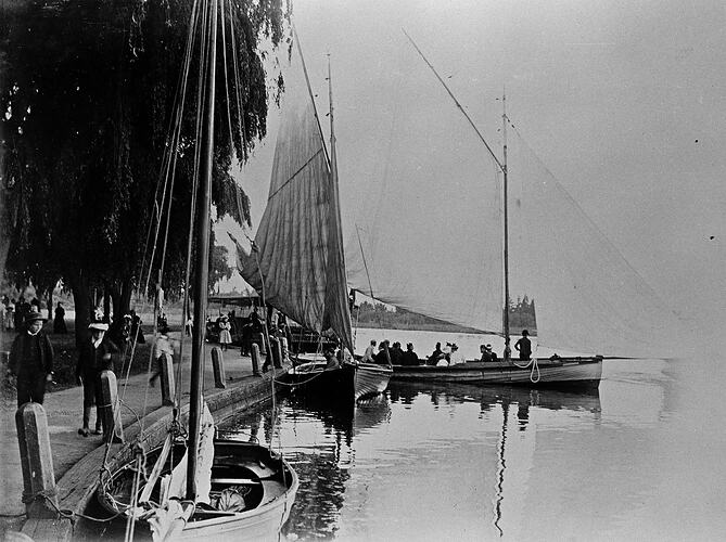 People walking along a promenade near moored sailing boats.