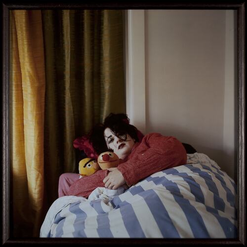 Woman wearing heavy face make-up lays in bed with blue and white striped quilt. She holds two stuffed toys.