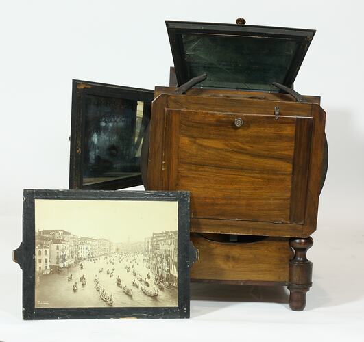 Polished wooden box-like object with viewing aperture and large square lens at front, several hinged doors.