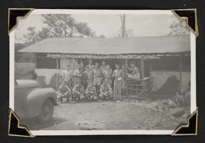 Group portrait of fifteen men standing in front of single story building.