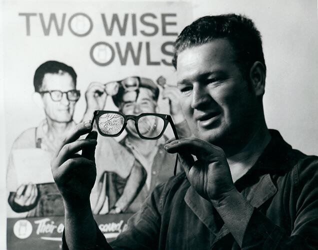 Man holding a broken pair of safety glasses in front of poster.