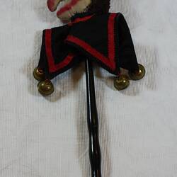 Mock sceptre with small jester's head made of cream cloth mounted on black wooden stick.