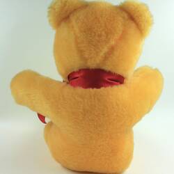 Back view of plush yellow teddy bear with red ribbon around its neck.