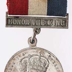 Round silver coloured medal with profile of crowned man and woman with text surrounding, and coloured ribbon.