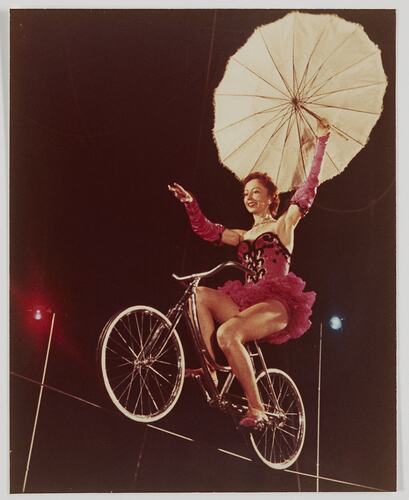 Woman riding a bicycle on a tightrope. She holds an umbrella.