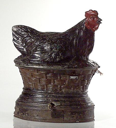 Metal hen painted black with red face, comb and wattles sits on a nest. Slot for coin and chute for eggs.