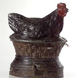 Metal hen painted black with red face, comb and wattles sits on a nest. Slot for coin and chute for eggs.