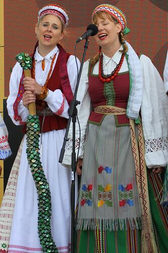 Two women in Lithuanian costume sing on stage. One holds a green wavy stick-shaped largerphone.