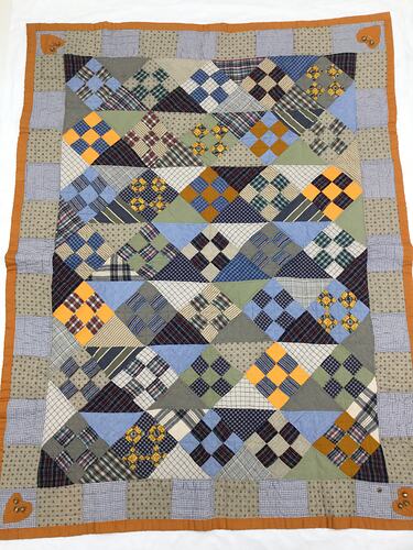 Colourful blue/grey patchwork quilt. Brown trim and heart in each corner.