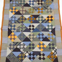 Colourful blue/grey patchwork quilt. Brown trim and heart in each corner.