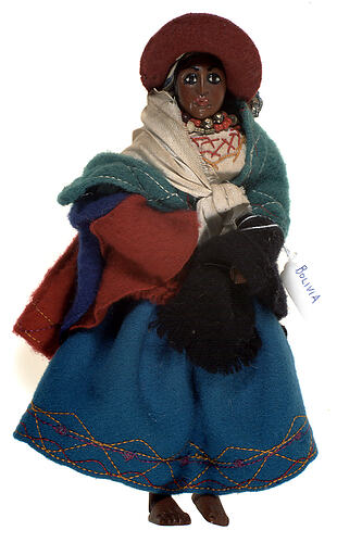 A doll wearing a blue skirt, a brown hat and three shawls.