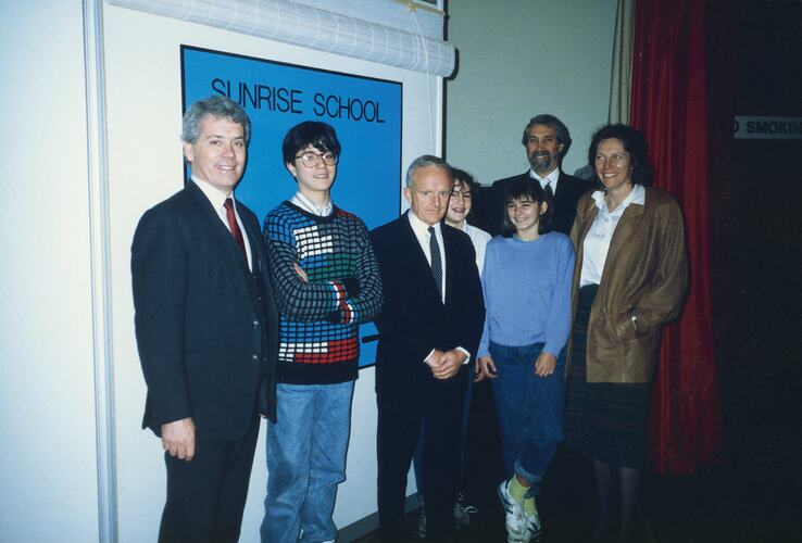 Group of adults and young people standing in front of a blue sign that says 'Sunrise School'.