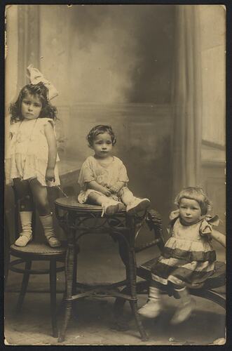 Three children pose wearing white. One stands on a chair, the middle sits on a table and the right child sits