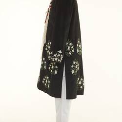Black coat with white floral detail over white chinos pants. Left profile.